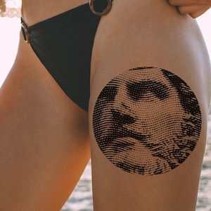 May include: A black bikini bottom with a brown ring detail. A black and white circular tattoo is on the wearer's thigh.