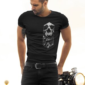 May include: A man wearing a black t-shirt with a white skull graphic stands next to a black motorcycle. The motorcycle has a black leather seat and a chrome headlight.