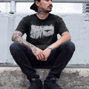 May include: A man wearing a black t-shirt with a white graphic of a forest and birds. He is wearing a black baseball cap and black jeans. He is sitting on a concrete ledge.