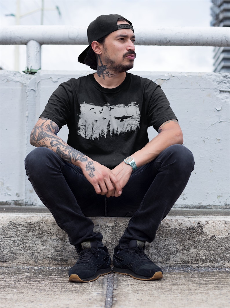 May include: A man wearing a black t-shirt with a white graphic of a forest and birds. He is wearing a black baseball cap and black jeans. He is sitting on a concrete ledge.