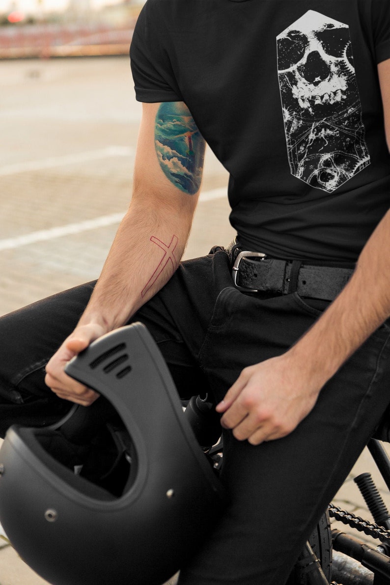 May include: A person wearing a black t-shirt with a white skull graphic. The person has a tattoo on their arm of a blue sky with clouds and a cross. The person is holding a black motorcycle helmet.