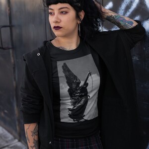 May include: A black t-shirt with a graphic print of a winged statue holding a sword. The statue is in a grayscale color scheme. The person wearing the shirt is standing in front of a metal wall.