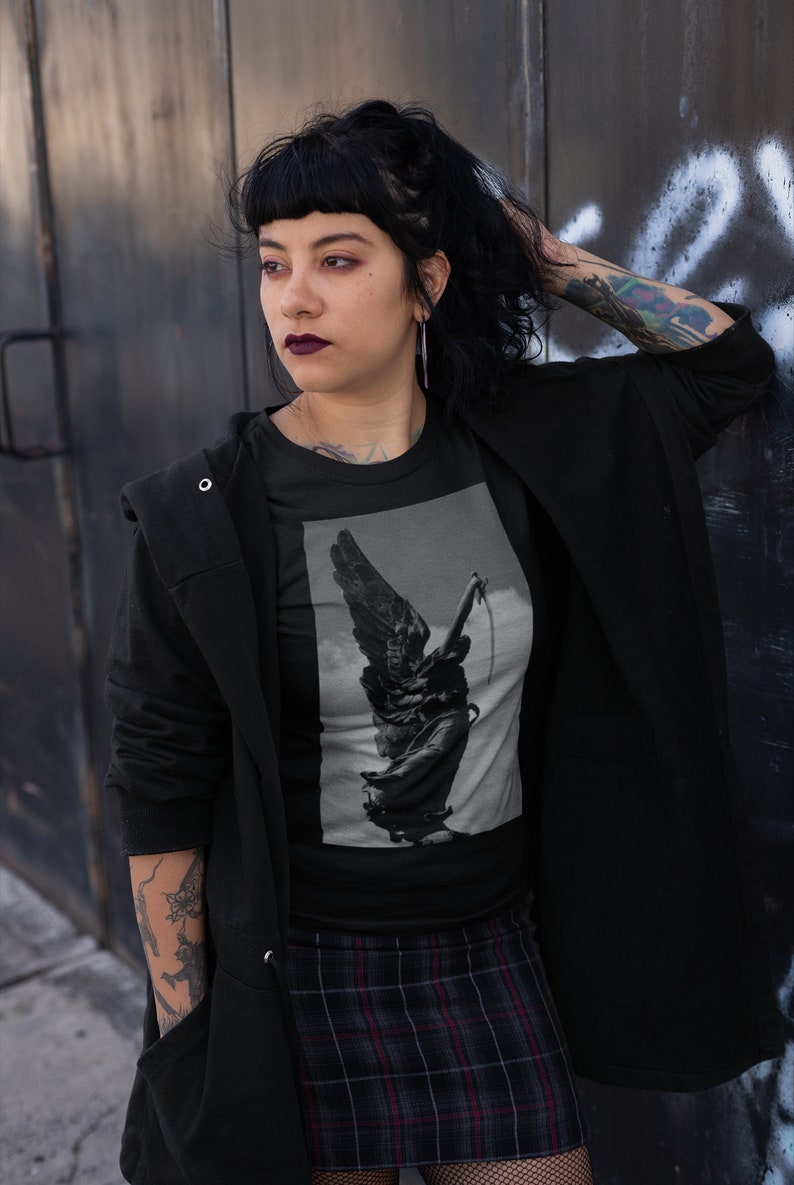 May include: A black t-shirt with a graphic print of a winged statue holding a sword. The statue is in a grayscale color scheme. The person wearing the shirt is standing in front of a metal wall.