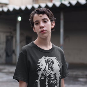 May include: A black t-shirt with a white graphic print of a skeletal figure in a hooded robe. The figure is standing with its arms crossed and has a distressed, grunge-style background.