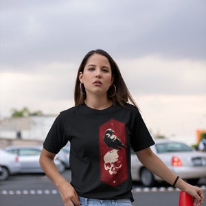 May include: A black t-shirt with a red and white graphic of a raven perched on a skull. The graphic is in a rectangular frame with rounded corners.