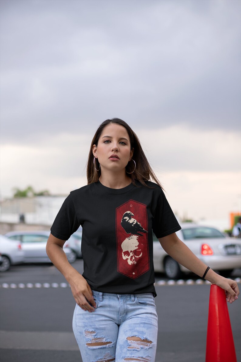 May include: A black t-shirt with a red and white graphic of a raven perched on a skull. The graphic is in a rectangular frame with rounded corners.