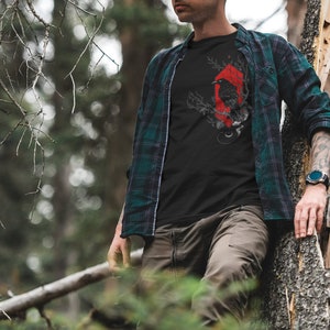May include: A man wearing a black t-shirt with a red and black crow graphic design. He is wearing a green and black plaid shirt over the t-shirt. He is standing in a forest and leaning against a tree.