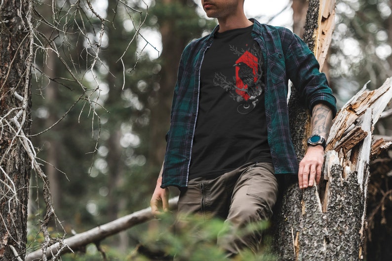 May include: A man wearing a black t-shirt with a red and black crow graphic design. He is wearing a green and black plaid shirt over the t-shirt. He is standing in a forest and leaning against a tree.