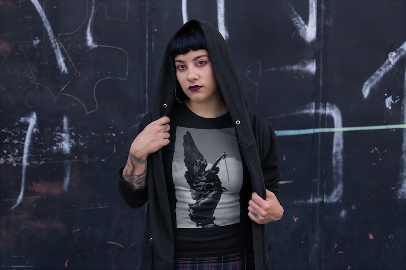 May include: A black hooded sweatshirt with a black t-shirt underneath. The t-shirt features a black and white image of an angel with wings holding a sword.