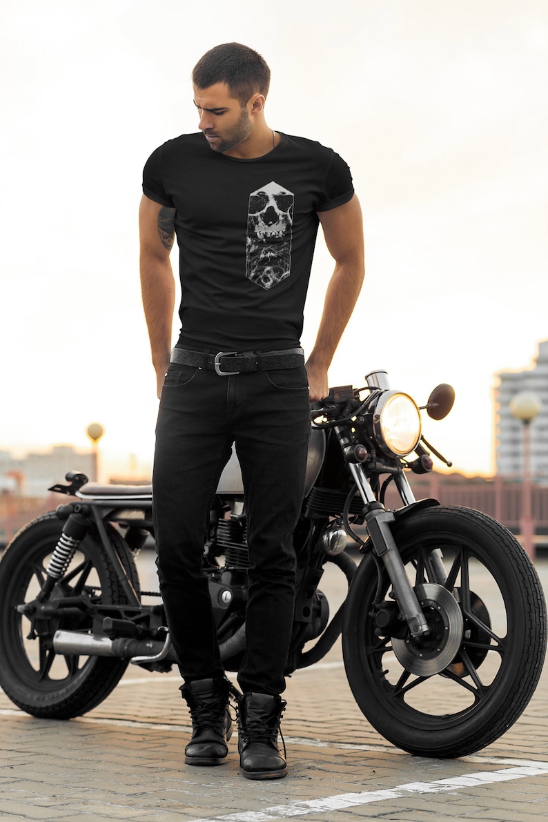 May include: A man wearing a black t-shirt with a white skull graphic stands next to a black motorcycle. The motorcycle has a black leather seat and a chrome headlight.