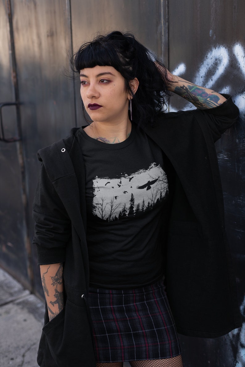 May include: A black t-shirt with a graphic print of a forest silhouette and a bird flying over it. The shirt is being worn by a person with black hair and a black jacket.