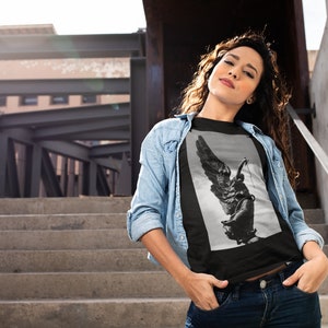 May include: A black t-shirt with a graphic print of a winged statue. The statue is in black and white and appears to be holding a sword. The t-shirt is being worn by a person standing on a set of stairs.