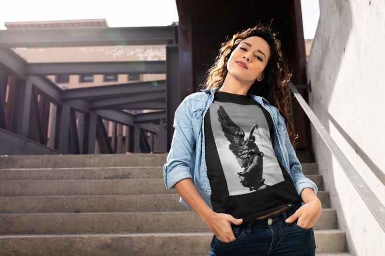 May include: A black t-shirt with a graphic print of a winged statue. The statue is in black and white and appears to be holding a sword. The t-shirt is being worn by a person standing on a set of stairs.