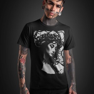 May include: A black t-shirt with a graphic print of a woman with snakes in her hair. The woman is looking down and has a sad expression on her face. The t-shirt is being worn by a person with tattoos on their arms.