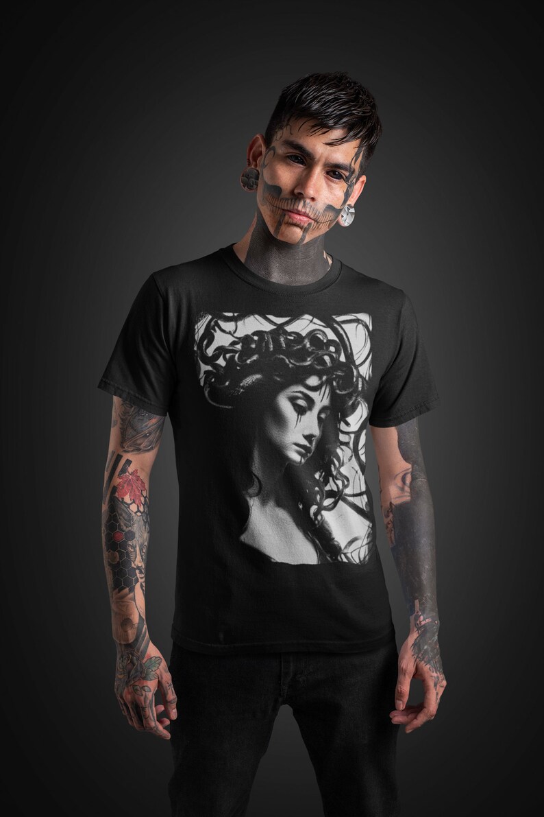 May include: A black t-shirt with a graphic print of a woman with snakes in her hair. The woman is looking down and has a sad expression on her face. The t-shirt is being worn by a person with tattoos on their arms.