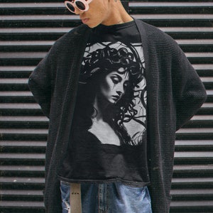 May include: A person wearing a black cardigan over a black t-shirt with a graphic print of a woman with snakes for hair. The person is wearing blue jeans with distressed details and white round sunglasses.