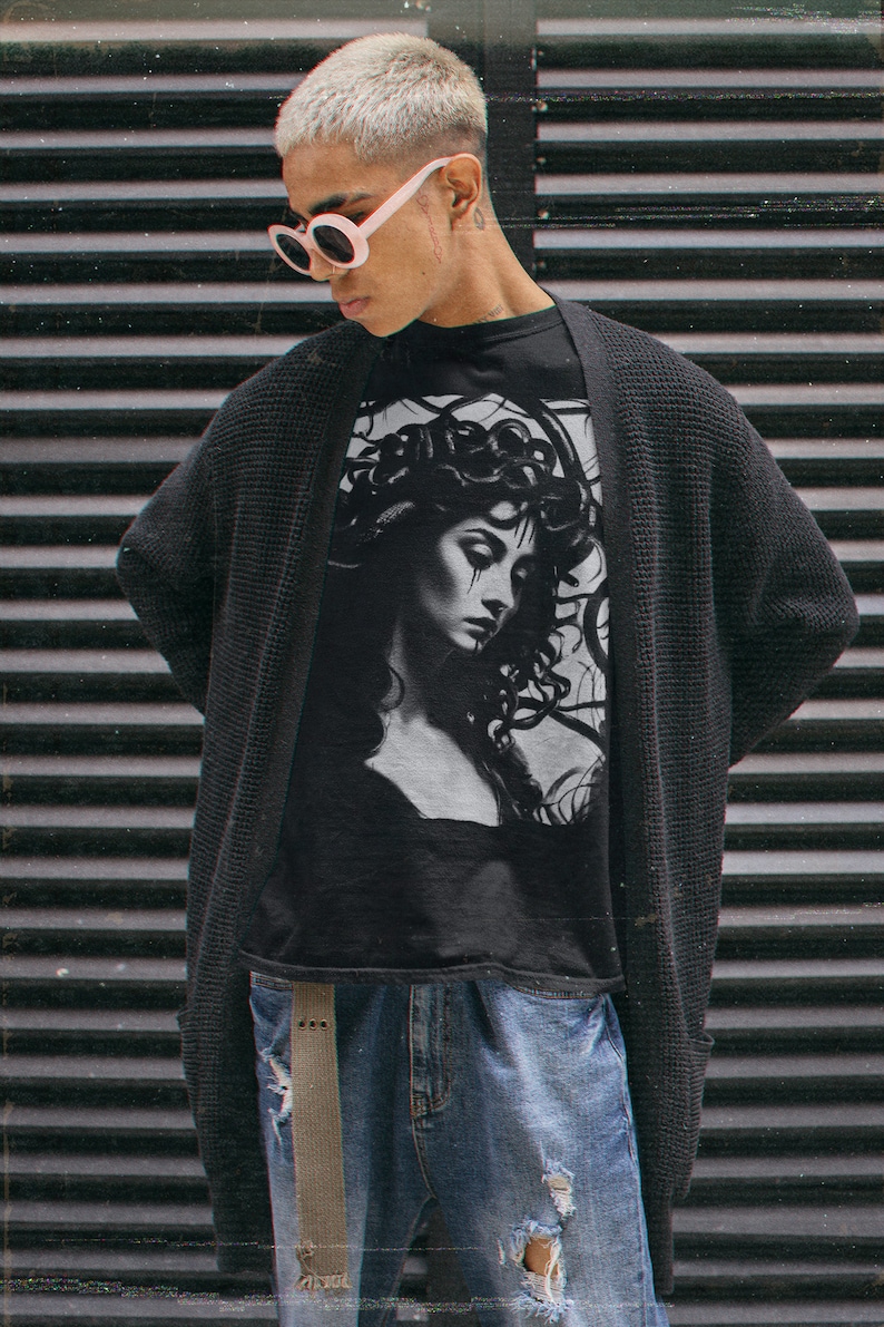 May include: A person wearing a black cardigan over a black t-shirt with a graphic print of a woman with snakes for hair. The person is wearing blue jeans with distressed details and white round sunglasses.