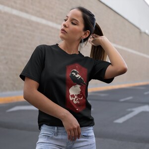 May include: Black t-shirt with a graphic of a crow perched on a skull. The graphic is in a red and white color scheme and is surrounded by a hexagonal frame.
