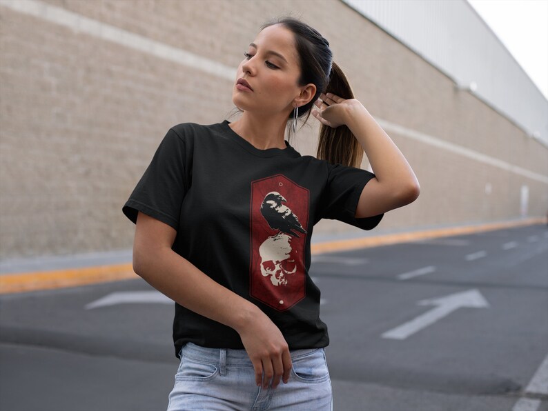 May include: Black t-shirt with a graphic of a crow perched on a skull. The graphic is in a red and white color scheme and is surrounded by a hexagonal frame.