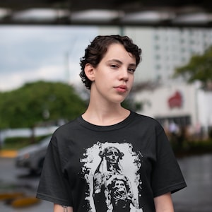 May include: A black t-shirt with a white graphic print of a woman in a hooded robe with a skull on her chest. The graphic has a distressed, vintage look.