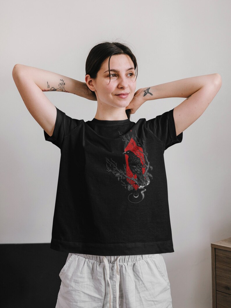 May include: Black t-shirt with a graphic print of a raven in a red diamond shape with branches and a crescent moon. The raven is perched on a branch with its wings spread.