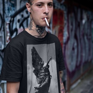 May include: A black t-shirt with a graphic print of a winged statue. The statue is in a black and white color scheme and appears to be holding a rope.