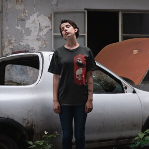May include: A person wearing a dark gray t-shirt with a black and white graphic of a skull and a bird. The person is standing in front of a white pickup truck with the hood open.