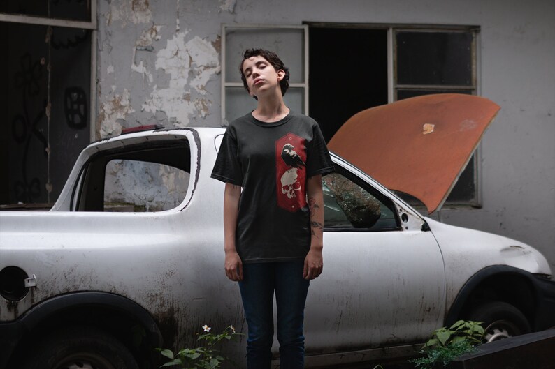 May include: A person wearing a dark gray t-shirt with a black and white graphic of a skull and a bird. The person is standing in front of a white pickup truck with the hood open.
