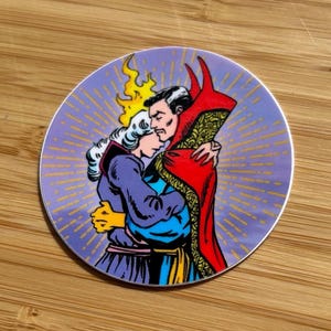 May include: A round sticker featuring a comic-style illustration of two figures embracing. One figure wears a red cloak and the other a purple outfit. The background is a light purple with radiating yellow lines.