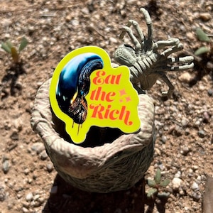 May include: A yellow sticker with the text "Eat the Rich" and an alien head graphic is placed on a textured, gray, egg-shaped object. A gray, alien-like creature is positioned behind the egg-shaped object. The scene is set outdoors on a bed of dirt and small plants.