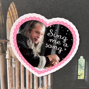 May include: Heart-shaped sticker featuring a portrait of a man with long gray hair and the text "Sing me a song." The sticker has a pink scalloped border and a black background with white stars.