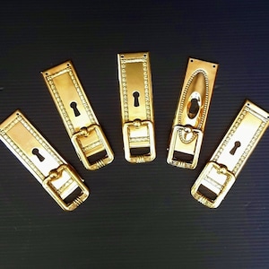 May include: Five gold-toned metal door hardware plates with keyholes and decorative edges. The plates have different shapes and sizes.
