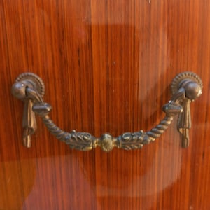 May include: Close-up of a wooden cabinet with a dark brown finish and a decorative metal handle. The handle is curved with ornate detailing and a bronze color, adding a vintage touch to the furniture.