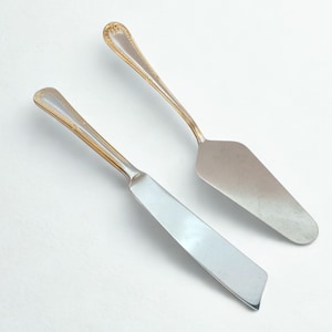 以下が含まれることがあります： 金色のアクセントが付いた2つの銀色のサービング用具。1つは、幅広の三角形の刃が付いたケーキサーバーです。もう1つは、長方形の刃が付いたバターナイフです。どちらも装飾的なハンドルが付いており、白い背景に配置されています。