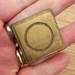 Puede incluir: Un objeto cuadrado de latón antiguo con un diseño circular en el centro. El objeto tiene una apariencia ligeramente desgastada, lo que sugiere antigüedad. Los bordes están biselados y el diseño general es simple y geométrico.