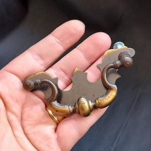 Op de afbeelding: Een messing kastgreep met een decoratief, sierlijk ontwerp. De greep heeft twee lussen en een centraal, plat, decoratief stuk.
