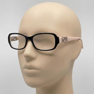May include: Rectangular spectacles featuring a black frame front and light pink temples. The temples are accented with a silver-toned metallic Greek key motif embedded with small clear crystals at the hinge connection point.