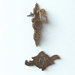 Puede incluir: Tirador de cajón y bocallave de latón antiguo. El tirador de cajón presenta un diseño decorativo con un tornillo para el montaje. La bocallave tiene un diseño ornamentado y curvado. Ambas piezas son de latón.