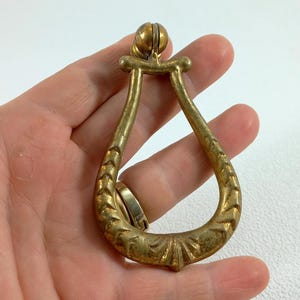 Op de afbeelding: Een vintage messing trekker met een druppelvorm. De trekker heeft een decoratief bloemmotief langs de gebogen randen en een kleine, ronde knop aan de bovenkant. Het metaal heeft een warme, verouderde patina.