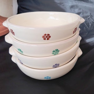 May include: A stack of four white ceramic bowls with a scalloped rim. Each bowl has a different colored floral pattern: red, green, blue, and purple.