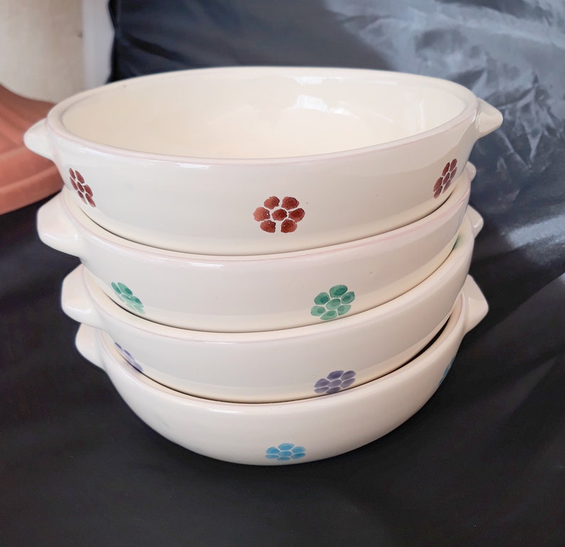 May include: A stack of four white ceramic bowls with a scalloped rim. Each bowl has a different colored floral pattern: red, green, blue, and purple.