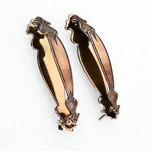 Puede incluir: Dos tiradores de puerta antiguos de color bronce con diseños ornamentados y detallados. Cada tirador tiene una forma ovalada alargada con elementos decorativos en la parte superior e inferior. Sobre fondo blanco.