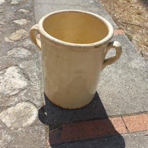 Op de afbeelding: Een cilindervormige, crèmekleurige keramische pot met een glanzende afwerking en twee kleine handvatten aan de zijkanten. De pot heeft een brede opening aan de bovenkant en een glad, afgerond lichaam met een egale, lichtgele glazuurlaag.