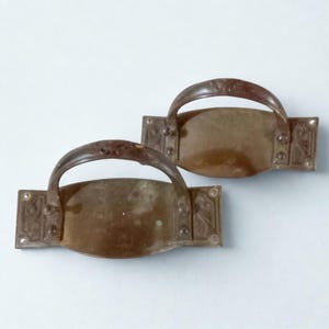 Puede incluir: Dos tiradores de cajón antiguos de bronce. Cada tirador tiene un asa curva y una placa trasera rectangular con detalles decorativos en las esquinas. El bronce tiene una pátina desgastada, lo que sugiere antigüedad. Estos tiradores son probablemente para muebles.