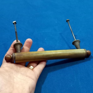 以下が含まれることがあります： 円筒形のハンドルと2つの取り付けポストを備えた、ヴィンテージの真鍮製引き出しプル。ハンドルは明るい茶色で、濃い茶色のエンドキャップが付いています。取り付けポストには装飾的なベースがあり、小さな正方形の頭が付いています。