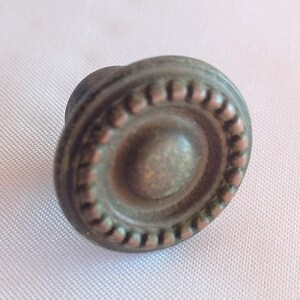 Op de afbeelding: Een ronde meubelknop met een donkerbronzen afwerking, voorzien van een verhoogde centrale koepel omringd door een textuurrand van kleine, gelijkmatig verdeelde kralen. Het beslag heeft een verweerde, metalen uitstraling met subtiele accenten.