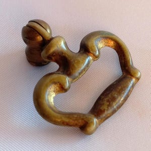 Puede incluir: Un tirador de cajón de metal en tono latón con un asa oscilante en forma de corazón unida a un poste de montaje circular. El herraje tiene un acabado envejecido con pátina visible y un diseño curvo y suave para agarrar y tirar.