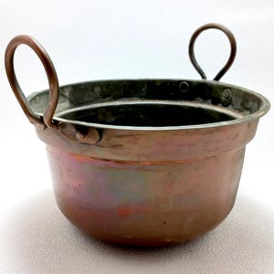 Pode incluir: Um pequeno pote de cobre antigo com duas alças arqueadas. O pote tem uma base arredondada e uma borda larga. O cobre tem um tom marrom-avermelhado quente com áreas de cor iridescente. As alças são presas às laterais do pote.