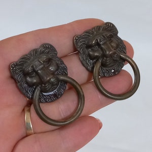 Puede incluir: Dos tiradores de cajones de bronce antiguo con forma de cabeza de león y asas de anillo. Las cabezas de león están detalladas con pelo y tienen un acabado desgastado.