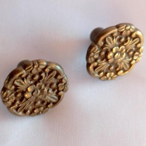 Op de afbeelding: Twee ronde metalen knoppen met een messing afwerking, elk voorzien van een gedetailleerd reliëf bloemmotief met verhoogde bloemblaadjes en bladeren. De knoppen hebben een cirkelvorm met een geschulpte rand en een centrale pin aan de achterzijde.